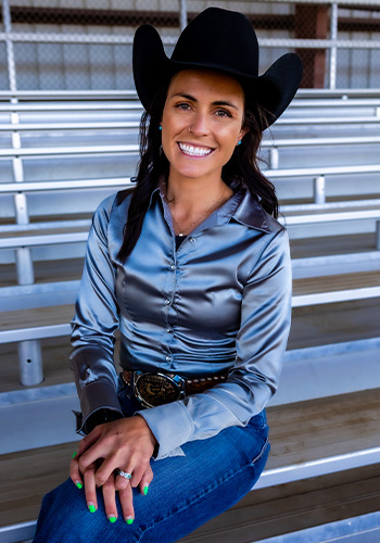 Sara Dobie smiles in jeans, a silky gray shirt, and black cowboy hat.